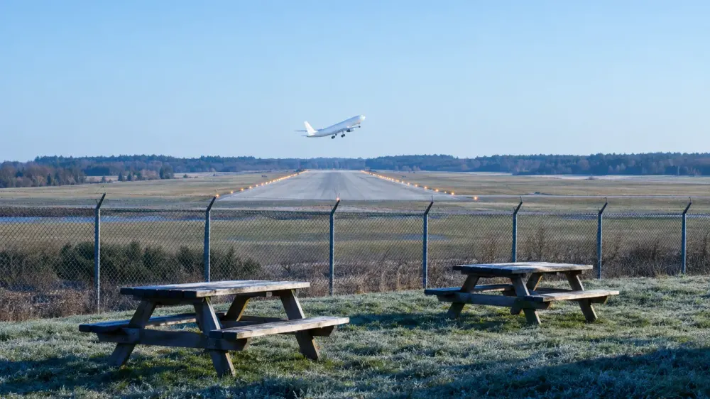 Two wooden picnic benches on a grass bank by a perimeter fence, a single runway beyond and a plain white airliner lifting off.