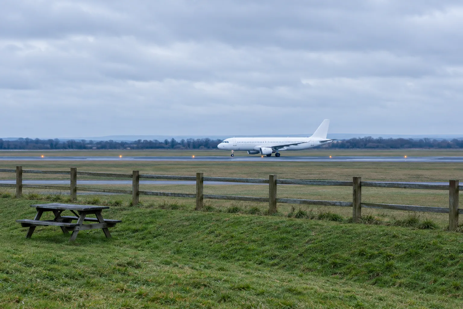 AI-made illustration of an overcast grass viewing area with a timber fence and an empty picnic bench, and a plain white twin-engine airliner on the runway in the distance.