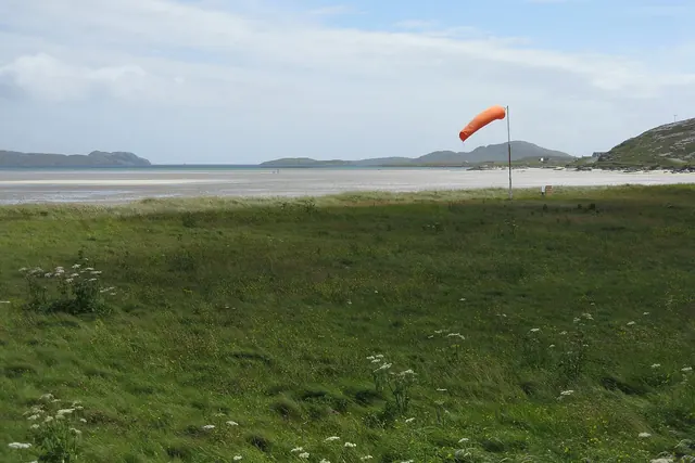 An orange windsock standing out in the breeze at Barra airfield, with the beach and hills behind.
