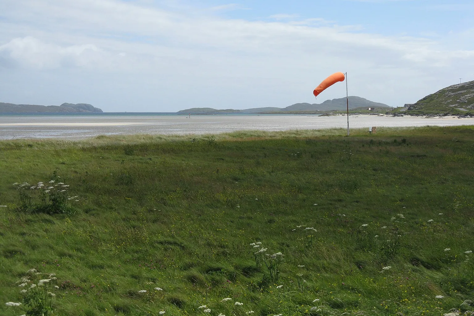 An orange windsock standing out in the breeze at Barra airfield, with the beach and hills behind.