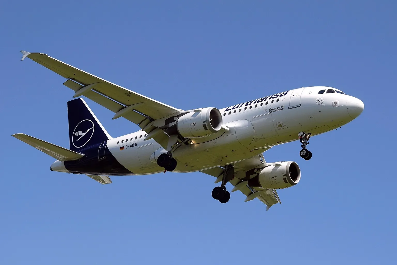 A Lufthansa Airbus A319 with its landing gear down, arriving at Birmingham Airport under a clear blue sky.