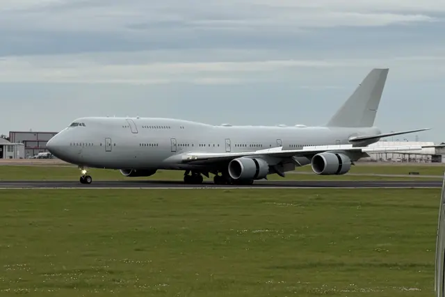 A plain grey Boeing 747 with no airline markings rolling along a runway, grass in the foreground.