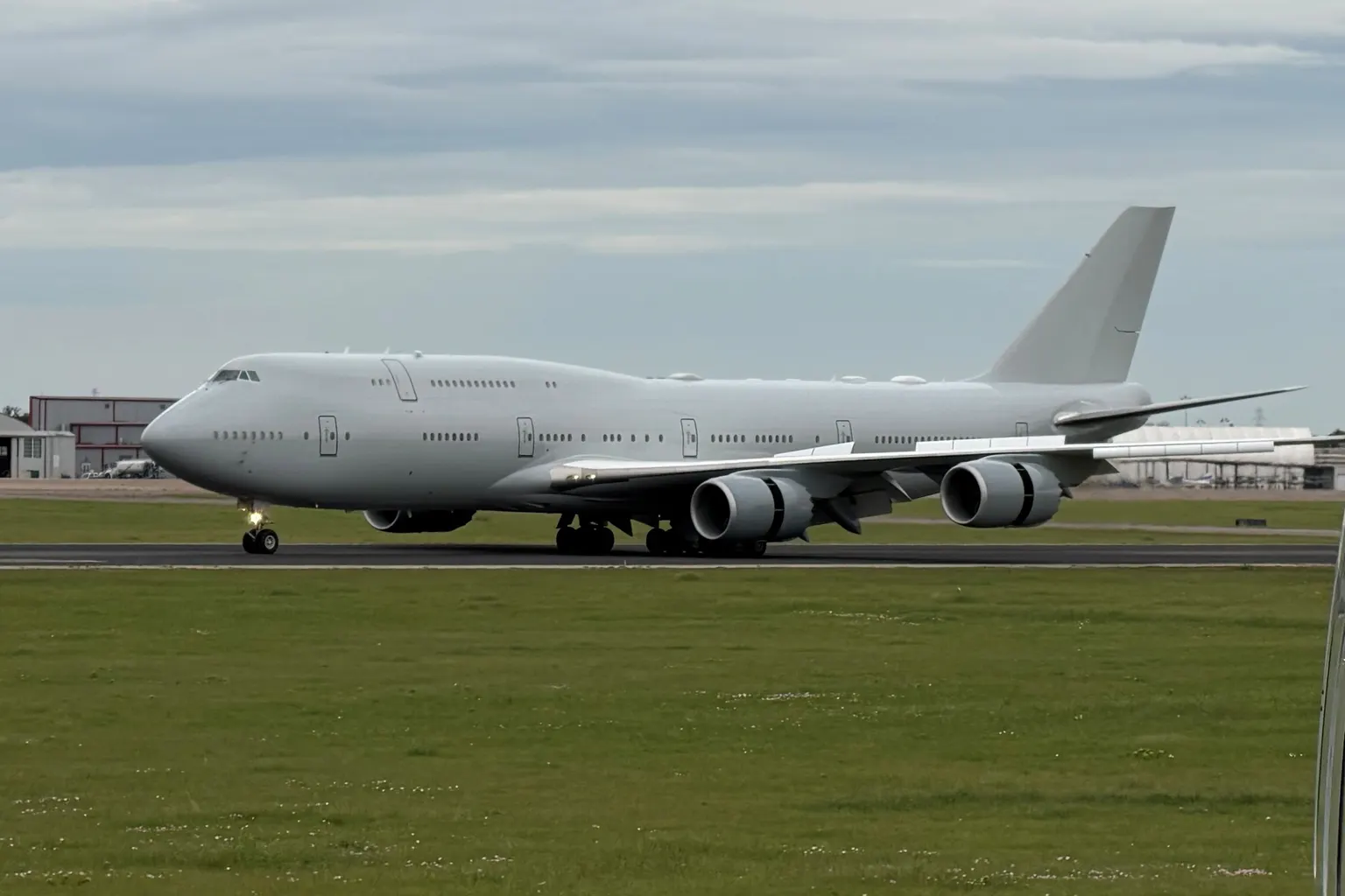 A plain white Boeing 747 with no markings on a runway, side on: fuselage, wings, four engines, tail and landing gear all in view.
