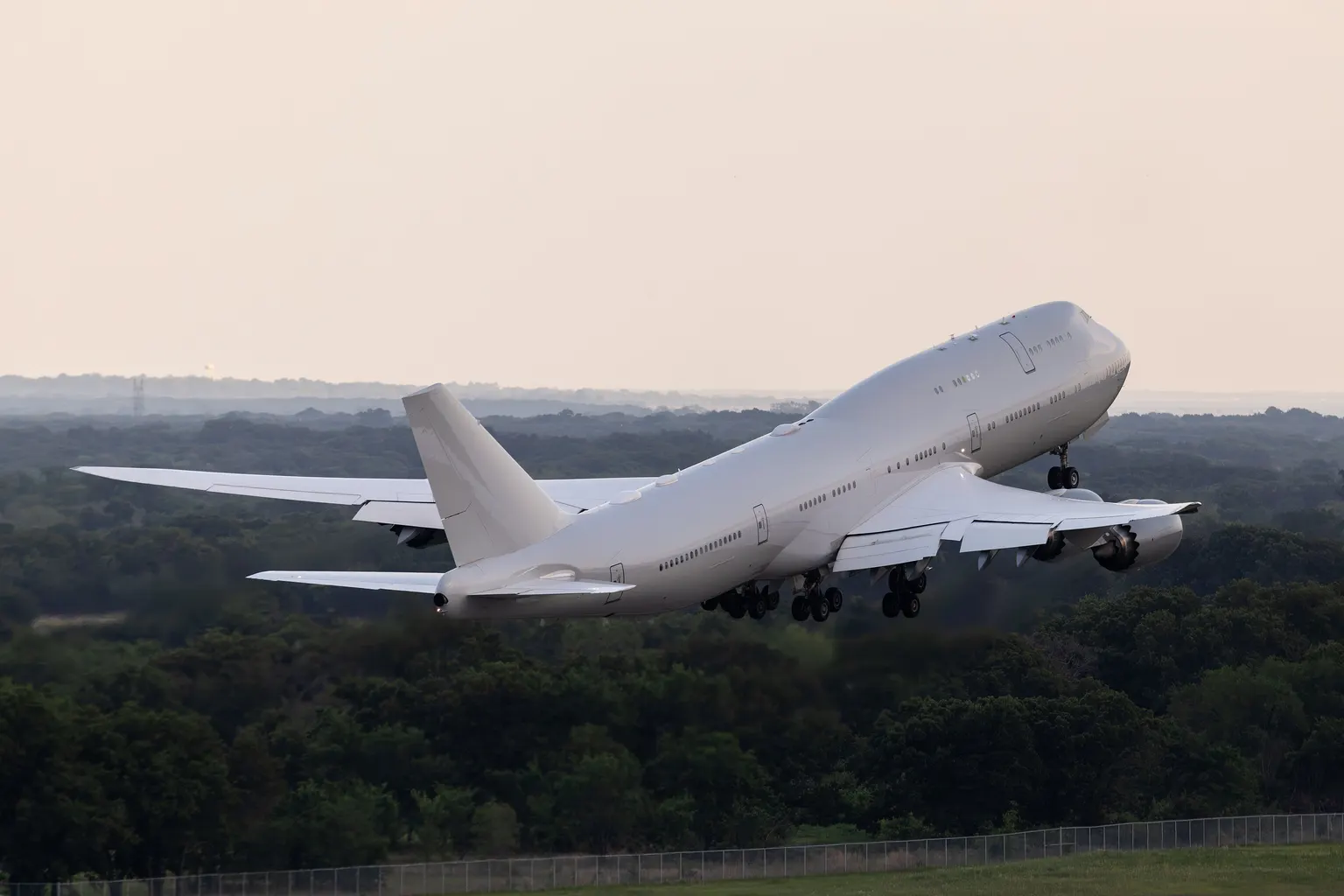 A plain white Boeing 747 with no airline markings climbing away over woodland in evening light.