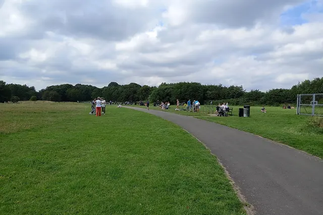 Plane spotters and picnickers on the grass beside a path at an airport viewing area, under a cloudy sky.