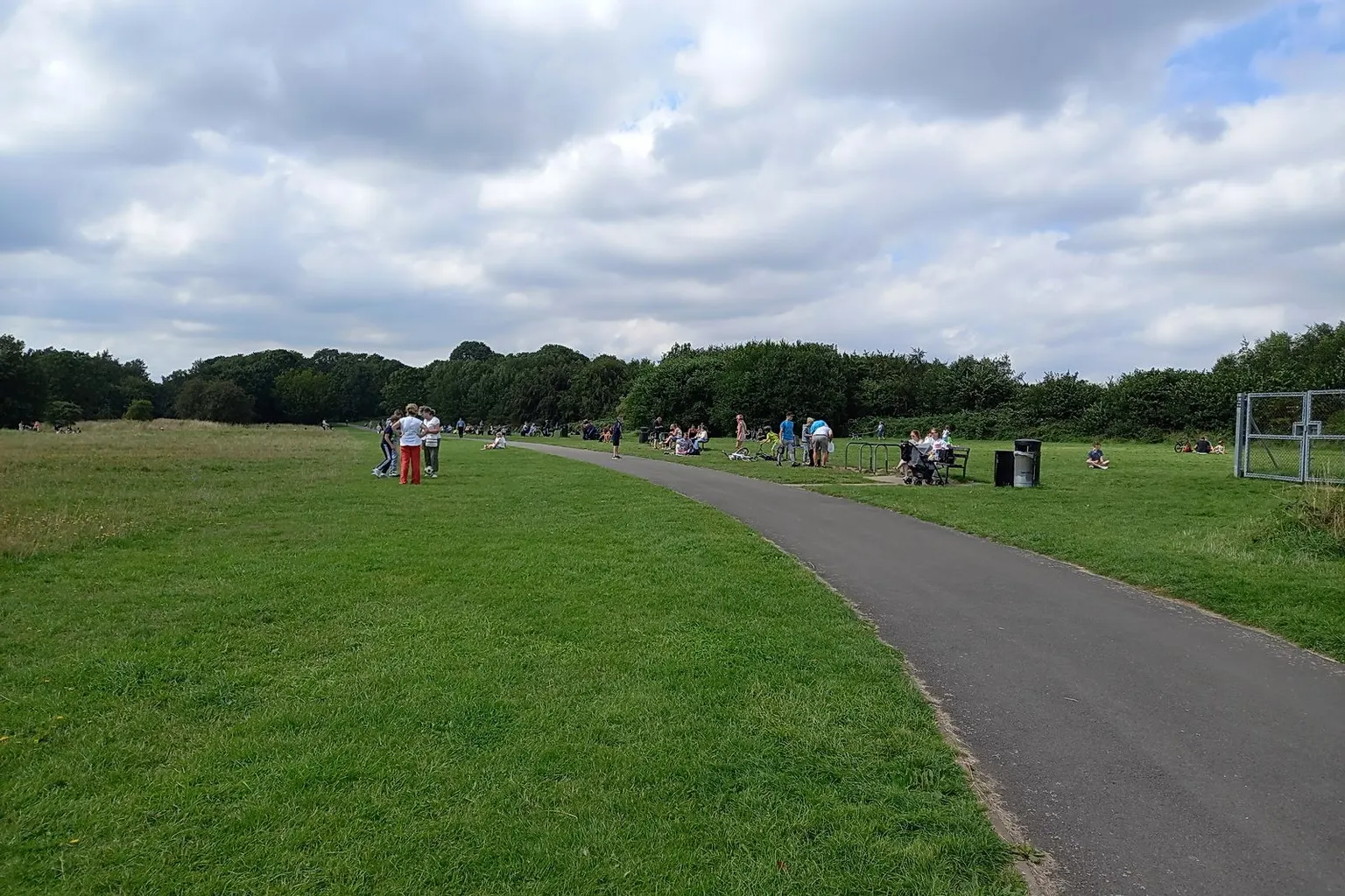 Plane spotters and picnickers on the grass at Sheldon Country Park, beside Birmingham Airport, under a summer sky.
