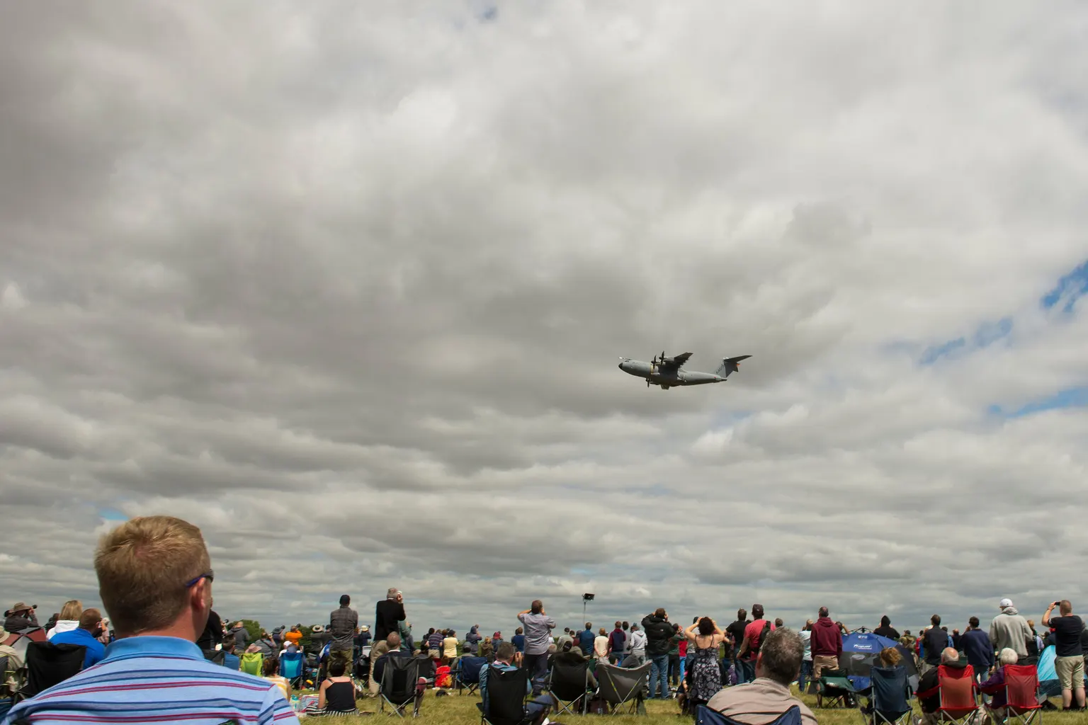 Spectators on the grass at the Royal International Air Tattoo, RAF Fairford, watching a grey transport aircraft fly past under heavy cloud.