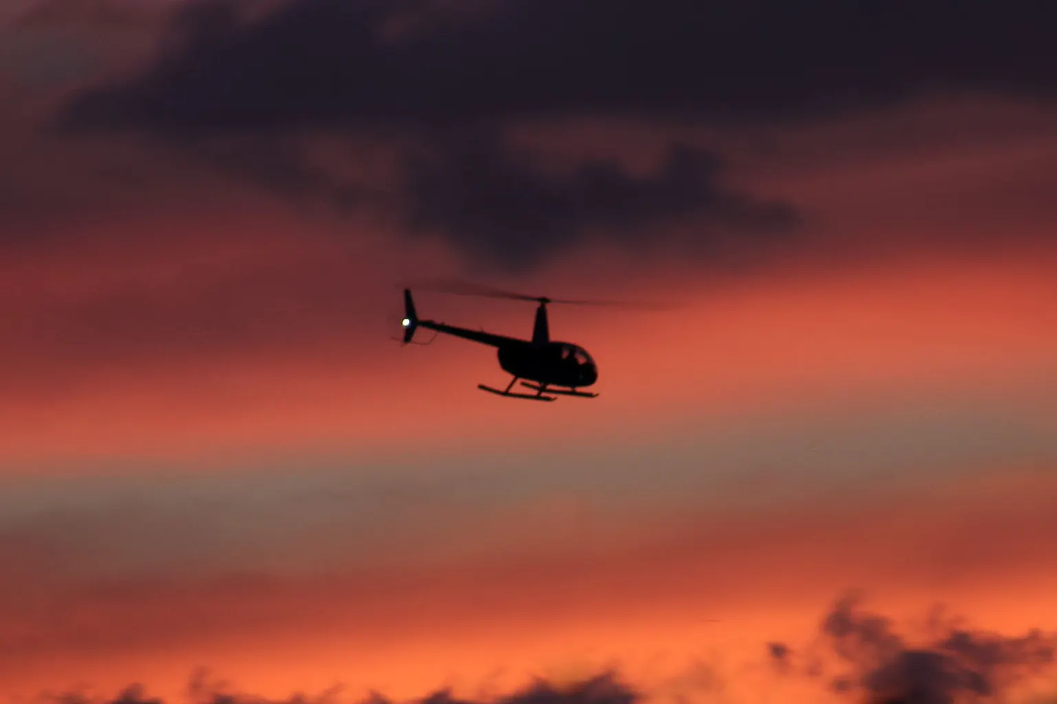 A light helicopter silhouetted against a red and orange sunset sky.