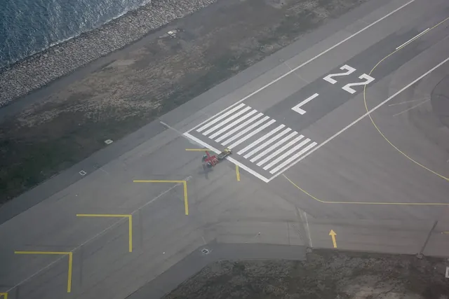 An aerial view of a runway threshold by the sea, the markings 22 and L painted beyond white stripes.