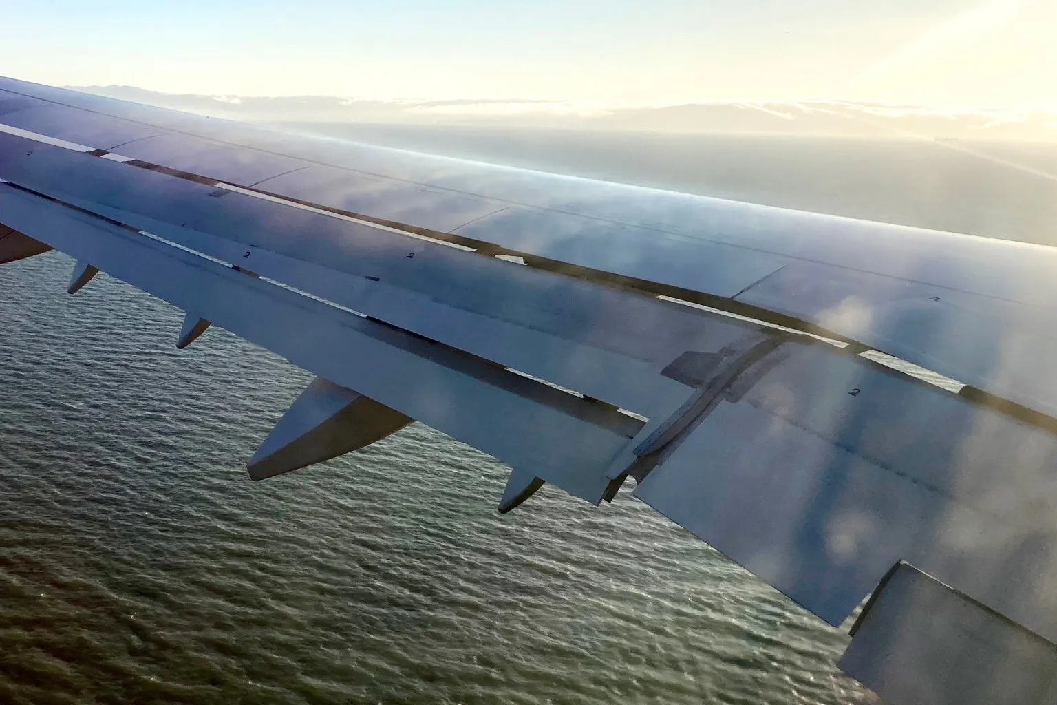 View from a cabin window over an airliner wing with its flaps lowered above the sea.