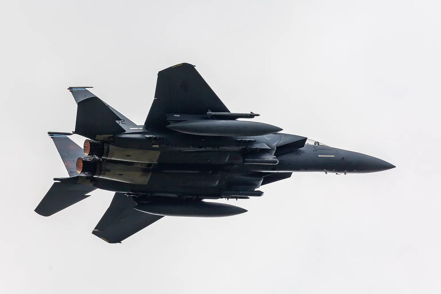 A US Air Force F-15E Strike Eagle from RAF Lakenheath banking overhead, seen from below against a pale sky.
