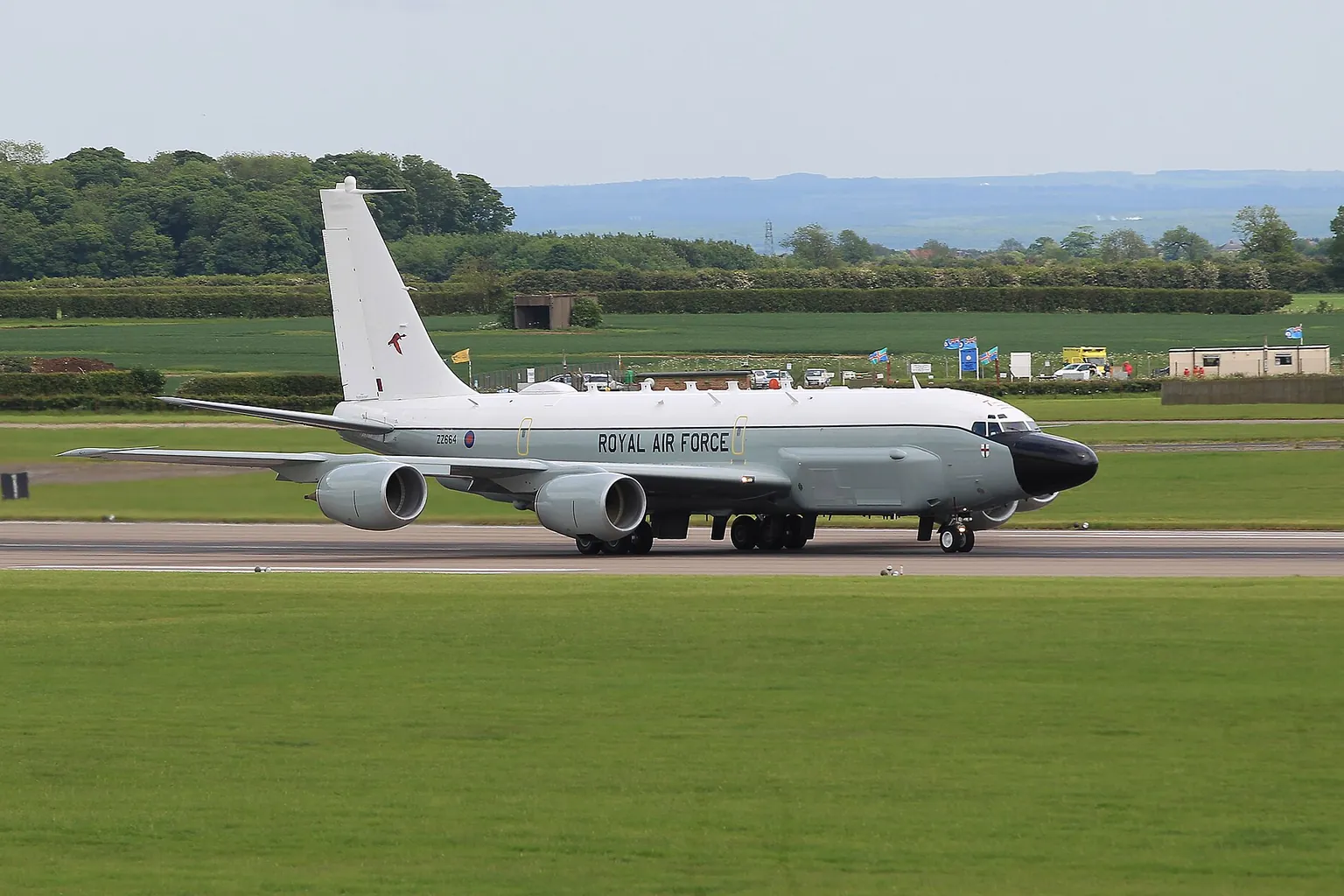 An RAF RC-135W Rivet Joint, white with a black nose, rolling along a runway between green fields.